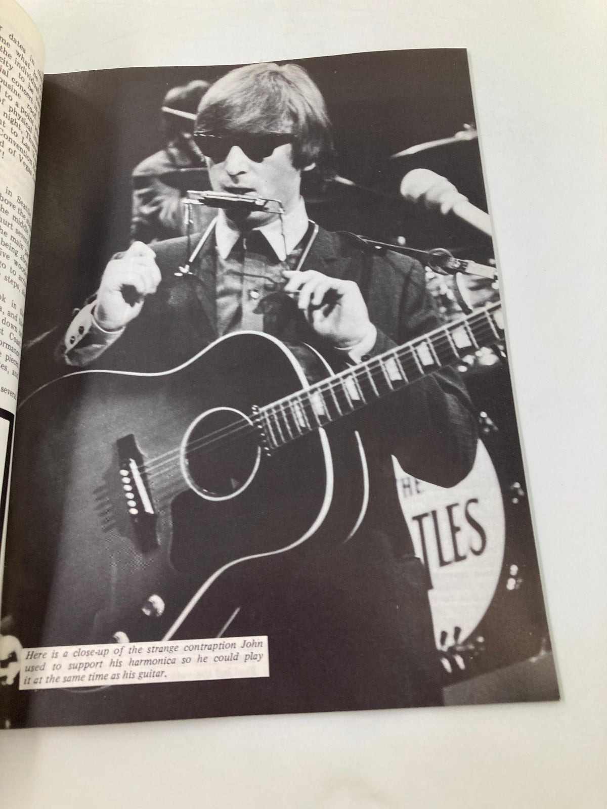 Close-up image in The Beatles Book Monthly Magazine October 1984 #102 of John Lennon playing harmonica while holding an acoustic guitar, showcasing his unique instrument setup.