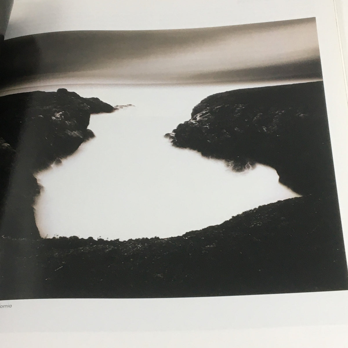 A striking black and white landscape photograph depicting a serene body of water surrounded by rugged cliffs, featured in Focus Photography Magazine February 2007, celebrating Ruth Bernhard's contributions to photography.