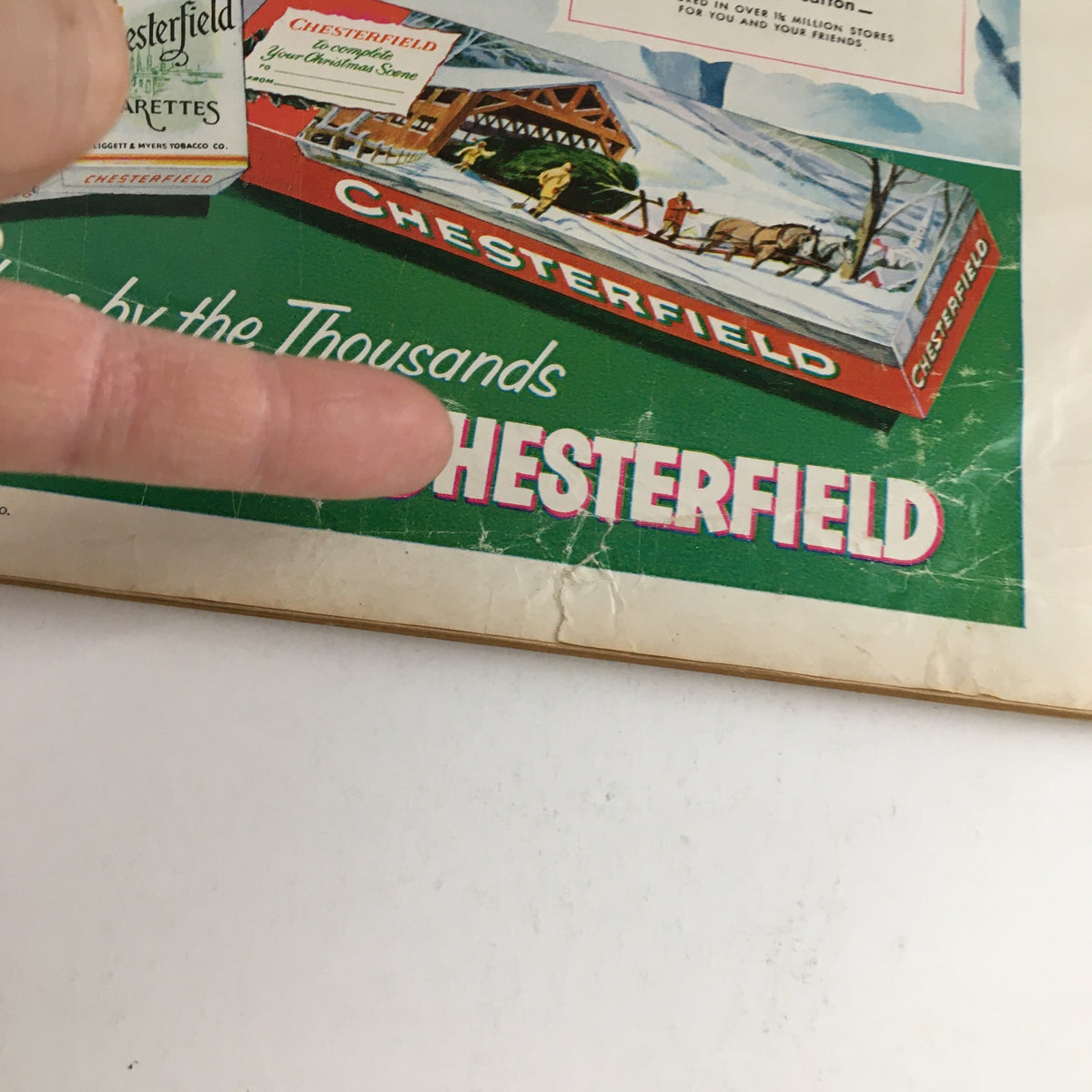 Close-up of the back cover of Photoplay Magazine December 1953 highlighting an advertisement for Chesterfield cigarettes with a finger pointing at the text. The ad includes a winter landscape theme.