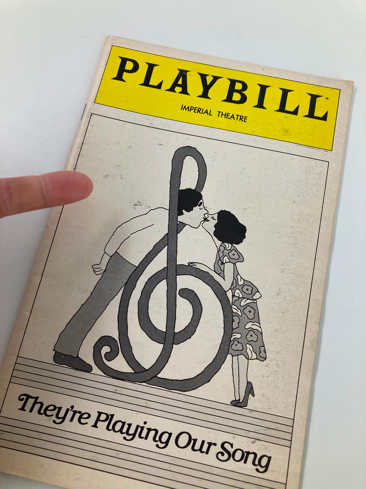 Close-up of the 1980 Playbill for 'They're Playing Our Song' showing a couple in a kiss pose next to a treble clef illustration.