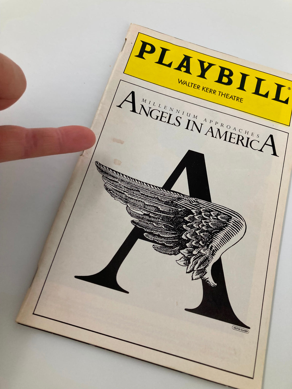 Close-up of the 1994 Playbill from Walter Kerr Theatre for "Angels in America," highlighting the winged 'A' design alongside a finger pointing to it, emphasizing its collectible nature.