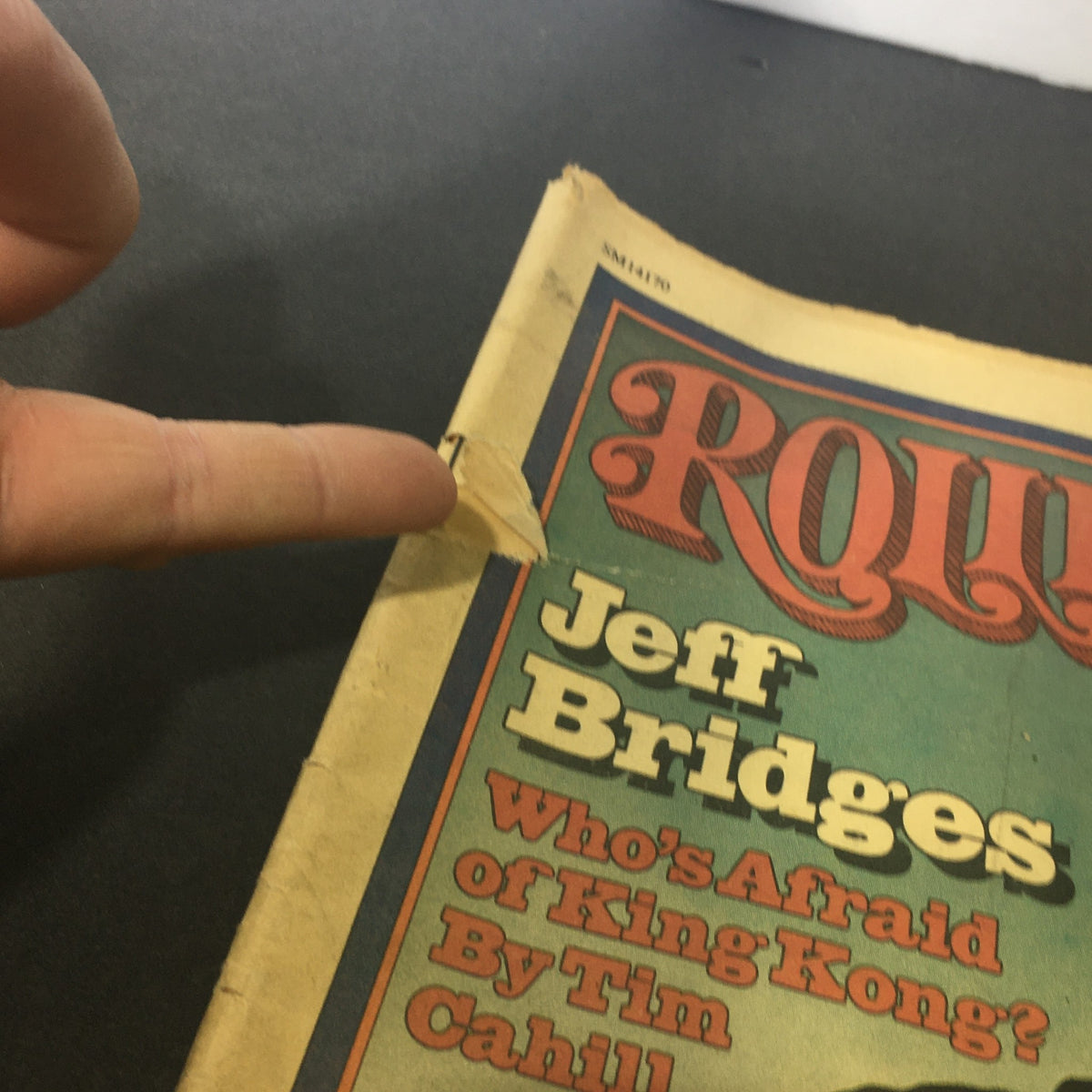 Close-up view of the front cover of the VTG Rolling Stone Magazine January 27 1977 featuring Jeff Bridges, showing wear with a visible tear and finger pointing to the damaged area.