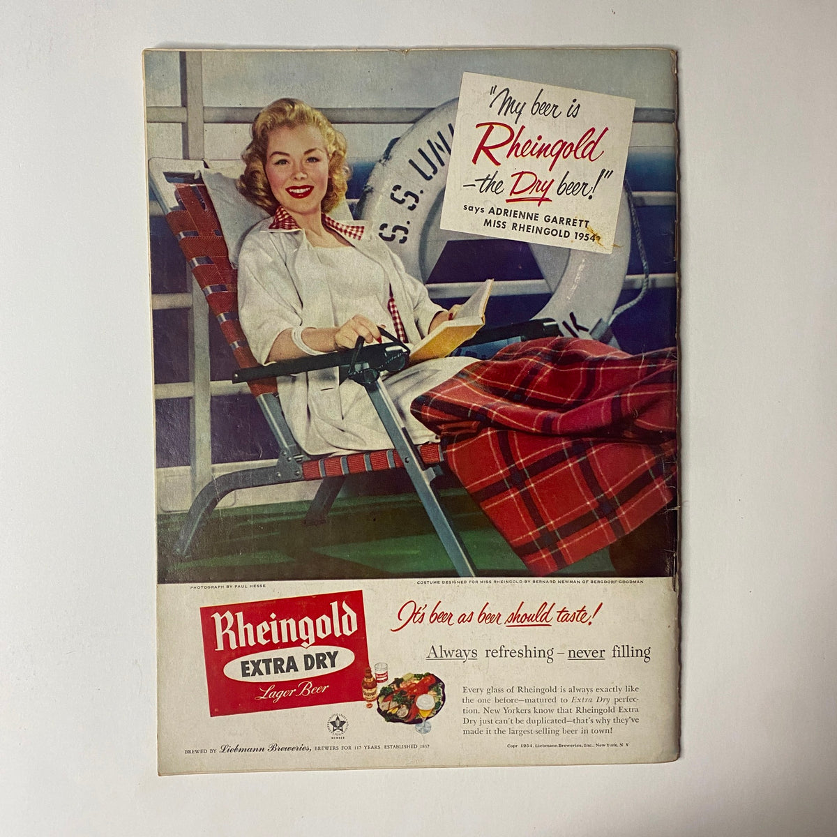 Vintage advertisement featuring a blonde woman relaxing on a deck chair, promoting Rheingold Extra Dry Lager Beer. The ad highlights the tagline, 'My beer is Rheingold, the Dry beer!' and features a signature from Adrienne Garrett, Miss Rheingold 1954. Perfect for collectors of mid-century beverage marketing.