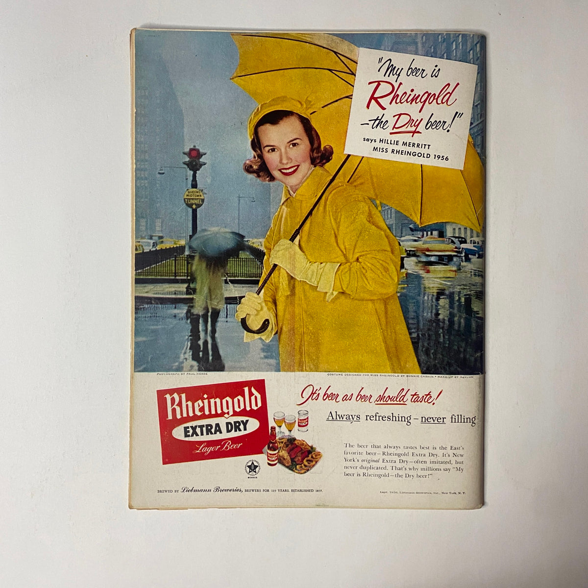 Back cover of Gourmet Magazine March 1956 featuring an advertisement for Rheingold Extra Dry Lager Beer with a woman in a yellow raincoat under an umbrella, promoting the brand's refreshing qualities.