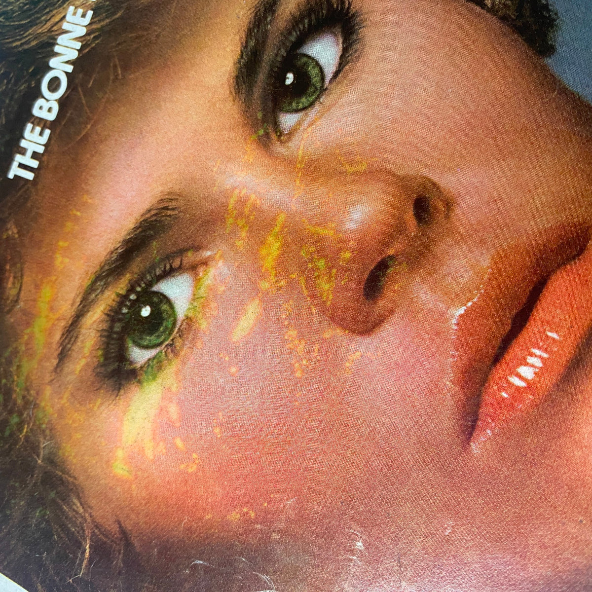 Detailed close-up of a model's face from a vintage Vogue Magazine July 1975 issue, displaying vibrant makeup and striking green eyes, capturing the essence of 1970s beauty trends.
