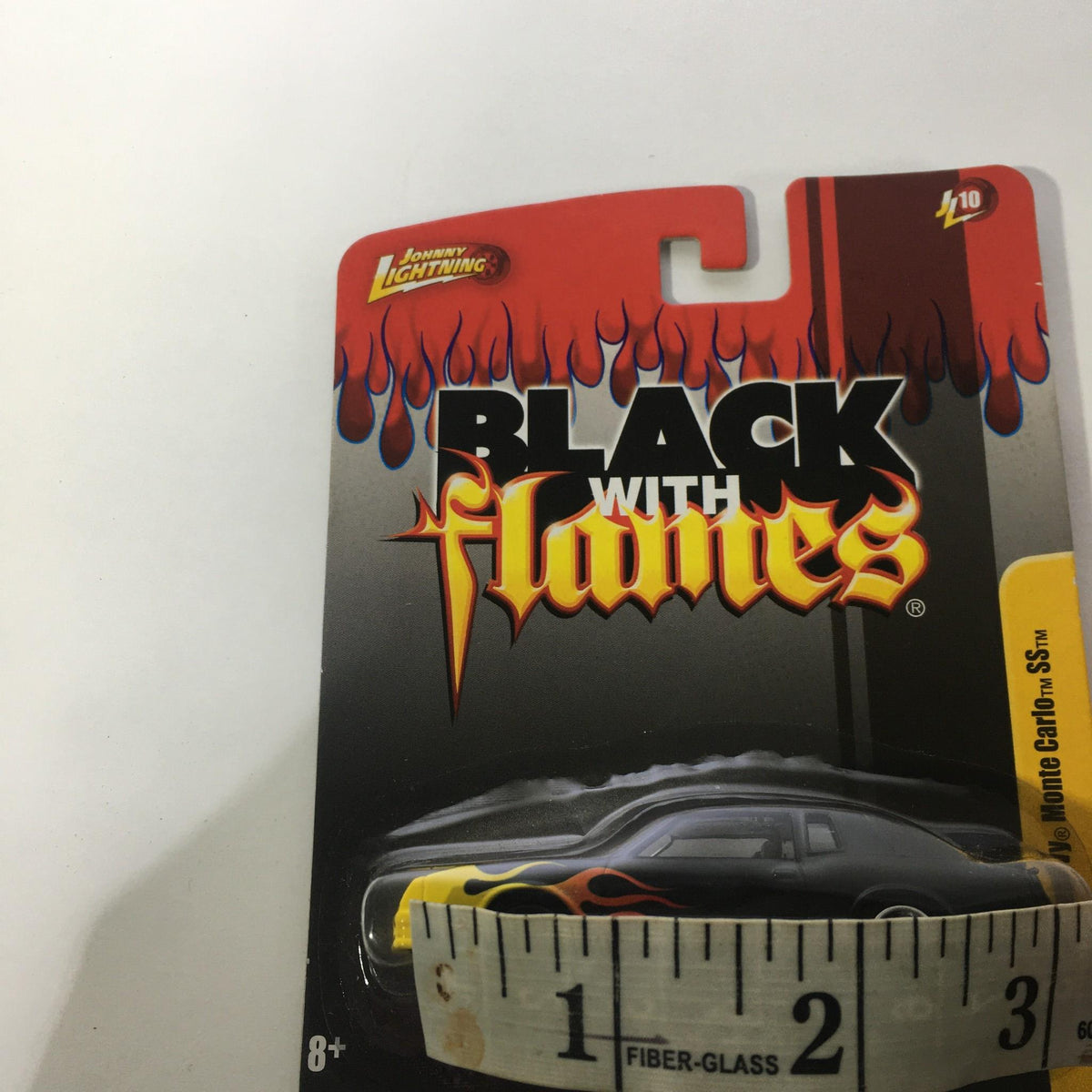 Front view of Johnny Lightning Black with Flames 1987 Chevy Monte Carlo SS packaging, featuring the design elements and a ruler for size reference.