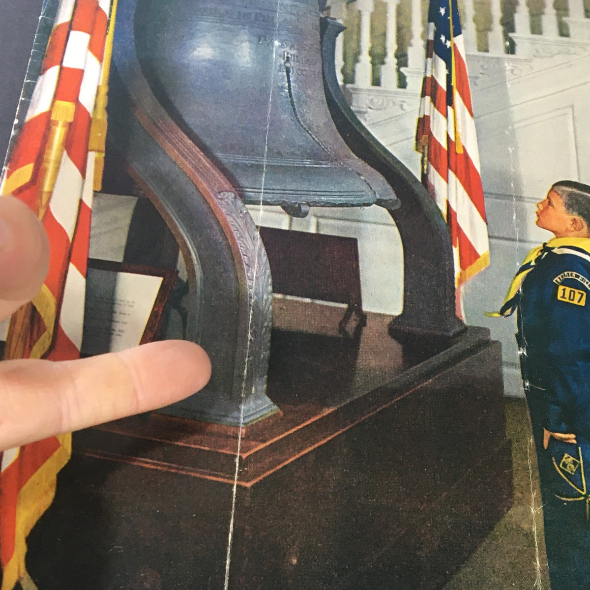 Close-up of a young boy in a sailor uniform observing a historical bell, accompanied by American flags, featured in the VTG These Times Magazine July 1953.