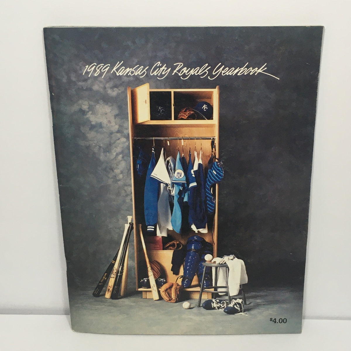 1989 MLB Official Team: Kansas City Royals Yearbook featuring a wooden locker filled with baseball uniforms, bats, and gear, showcasing the team's colors and memorabilia.