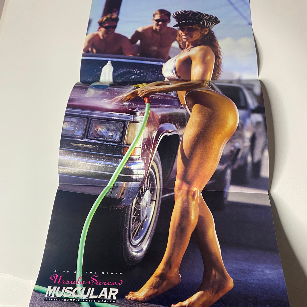 Fold-out poster from Muscular Development Magazine October 1996 featuring Ursula Sarcev posing sensually with a car and hose, emphasizing the glamour of bodybuilding.