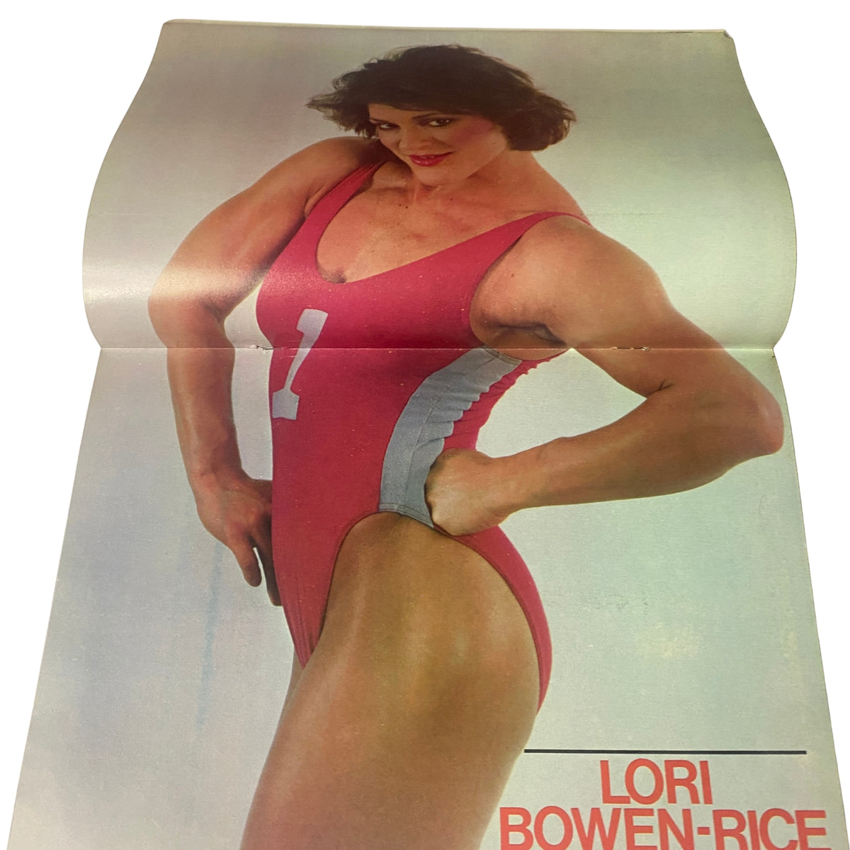 Lori Bowen-Rice featured in Muscle & Bodybuilder Magazine Fall 1985, showcasing her strong build in a red swimsuit with the number 1. This image is part of the magazine's attached color centerfold.