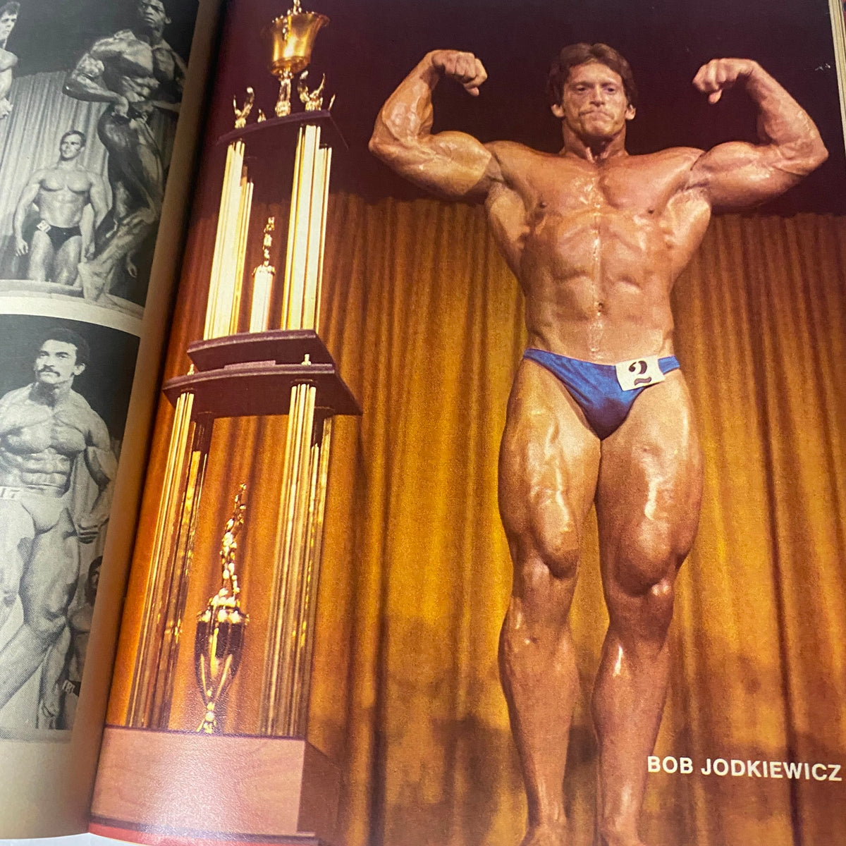 Bob Jodkiewicz posing at a bodybuilding competition, featured in Bodybuilder Magazine January 1980, Vol. 2 No. 2, with awards in the background.