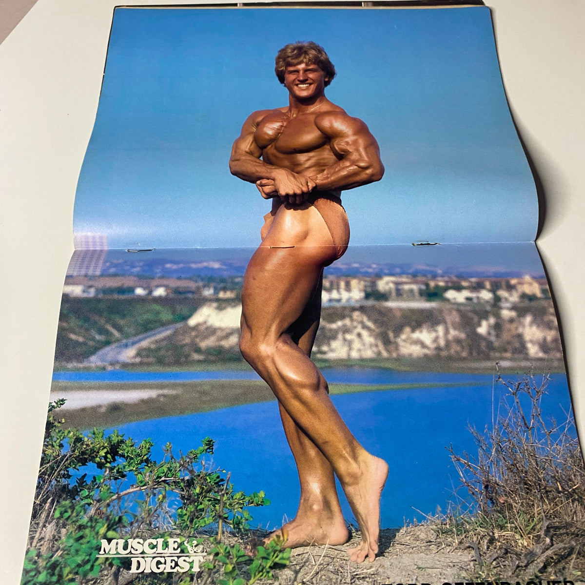 Centerfold from Muscle Digest Magazine April 1983 featuring a muscular male bodybuilder posing by a scenic water view.