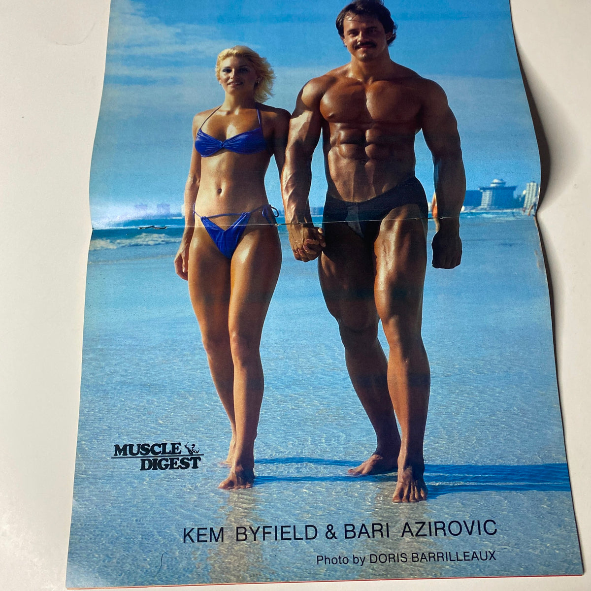 Muscle Digest Magazine December 1983 centerfold featuring models Kem Byfield and Bari Azirovic on the beach. Highlighting fitness and aesthetics in bodybuilding with a vibrant seaside backdrop.