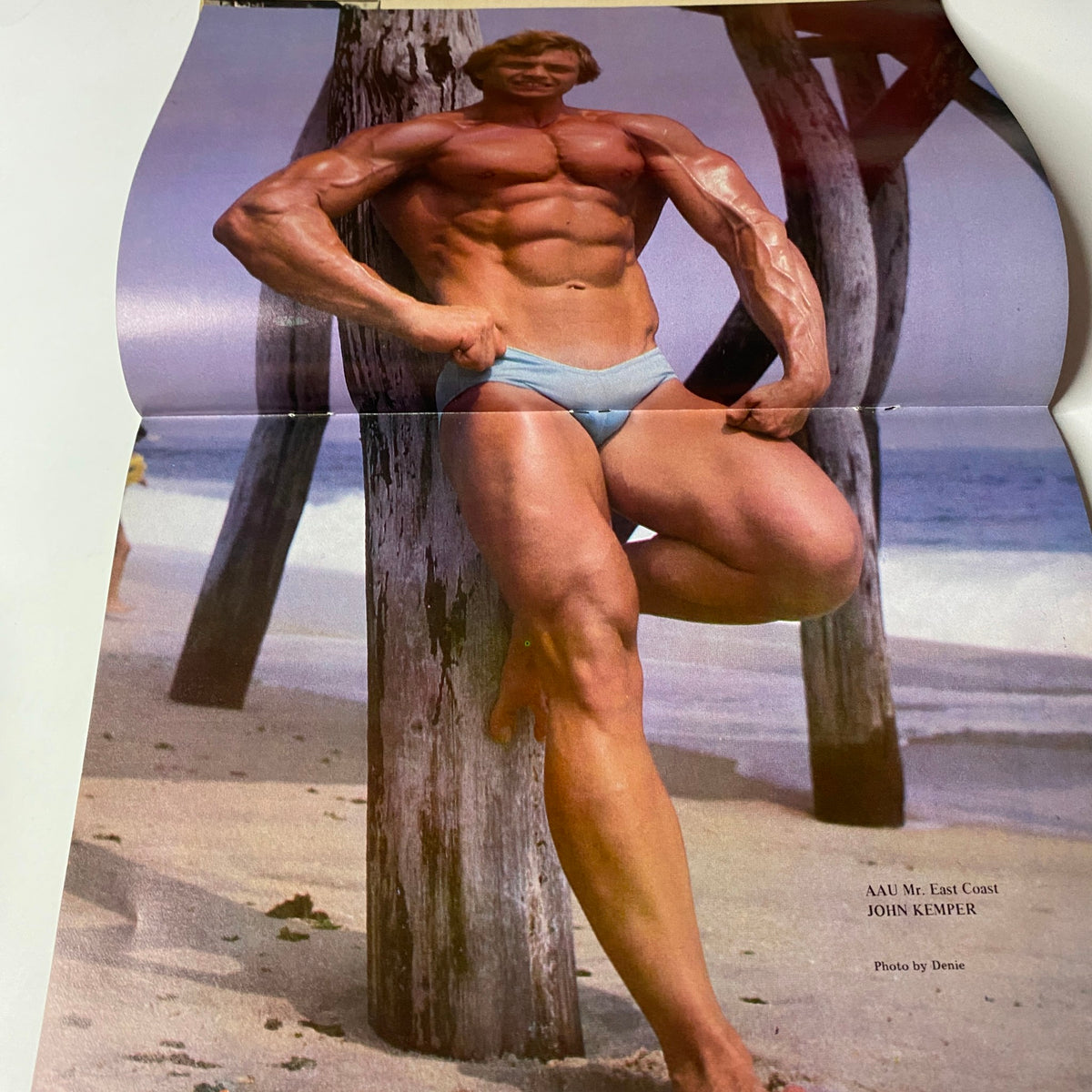 Muscle Training Illustrated Spring 1981, featuring a muscular male bodybuilder posing on the beach. This image showcases John Kemper, the AAU Mr. East Coast, highlighting the focus on strength training in the magazine.
