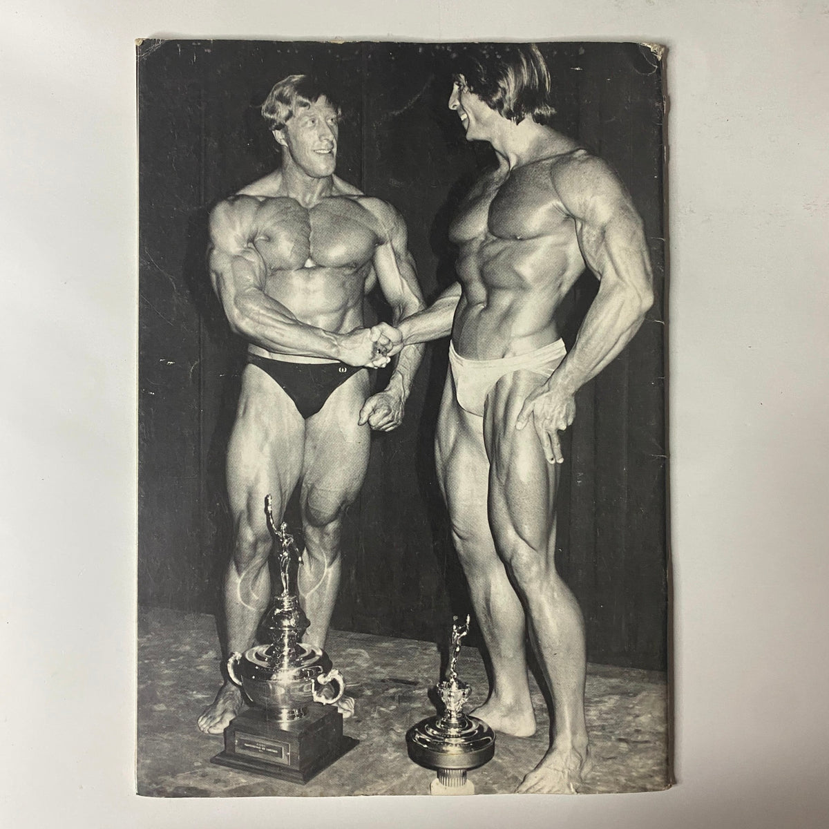 1977 NABBA Mr. Universe by Edward Hankey featuring two bodybuilders congratulating each other with their trophies, symbolizing achievement in the bodybuilding competition.