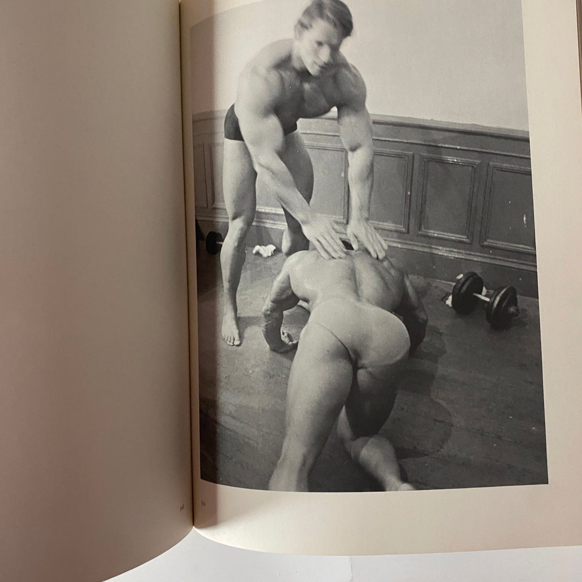 Black-and-white photograph from 'Pumping Iron: The Art & Sport of Bodybuilding', showcasing two bodybuilders engaged in a training session, emphasizing physical strength and camaraderie.