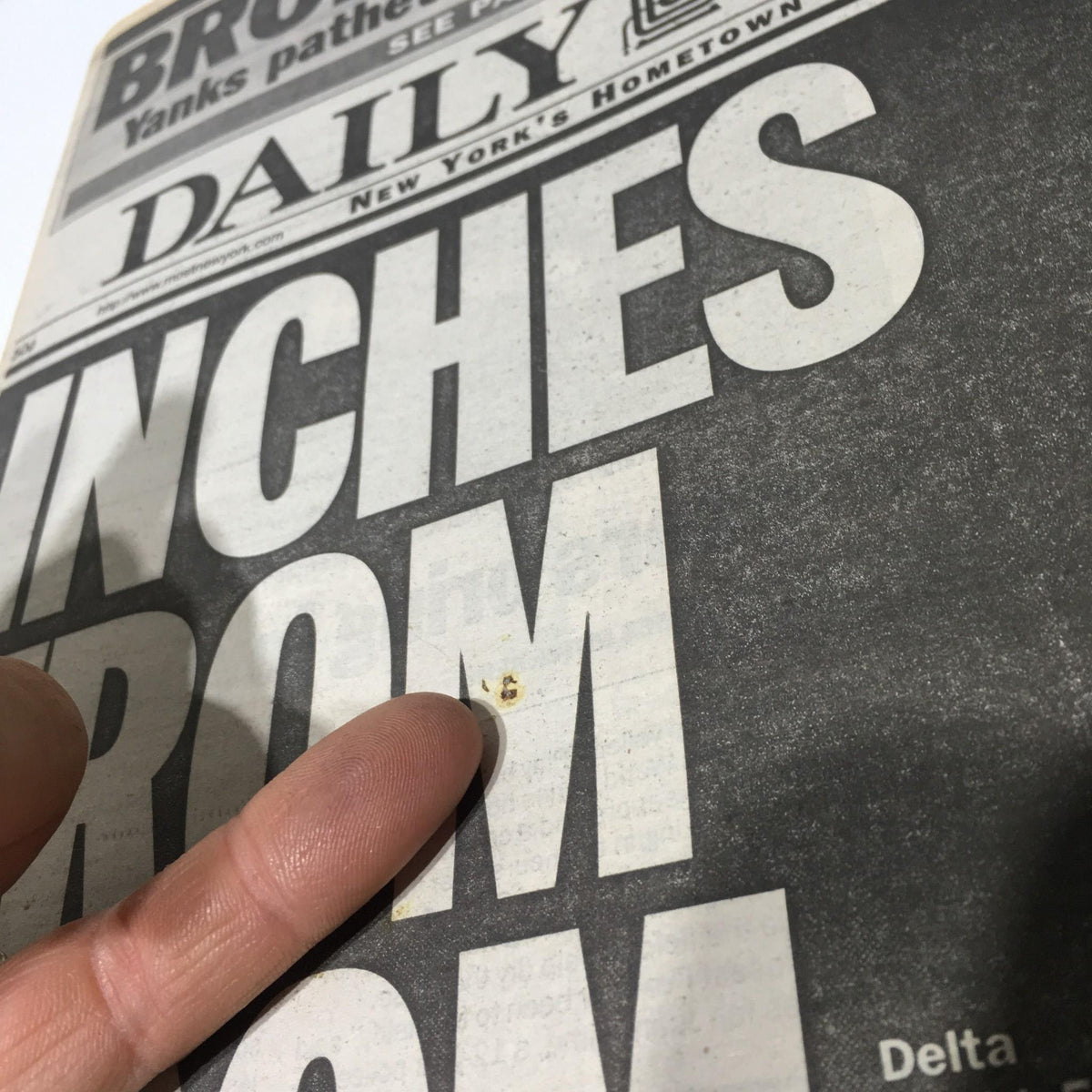 Close-up of 'Inches From Doom' headline from NY Daily News, showing a finger pointing at minor staining on the paper, highlighting its condition for potential buyers.