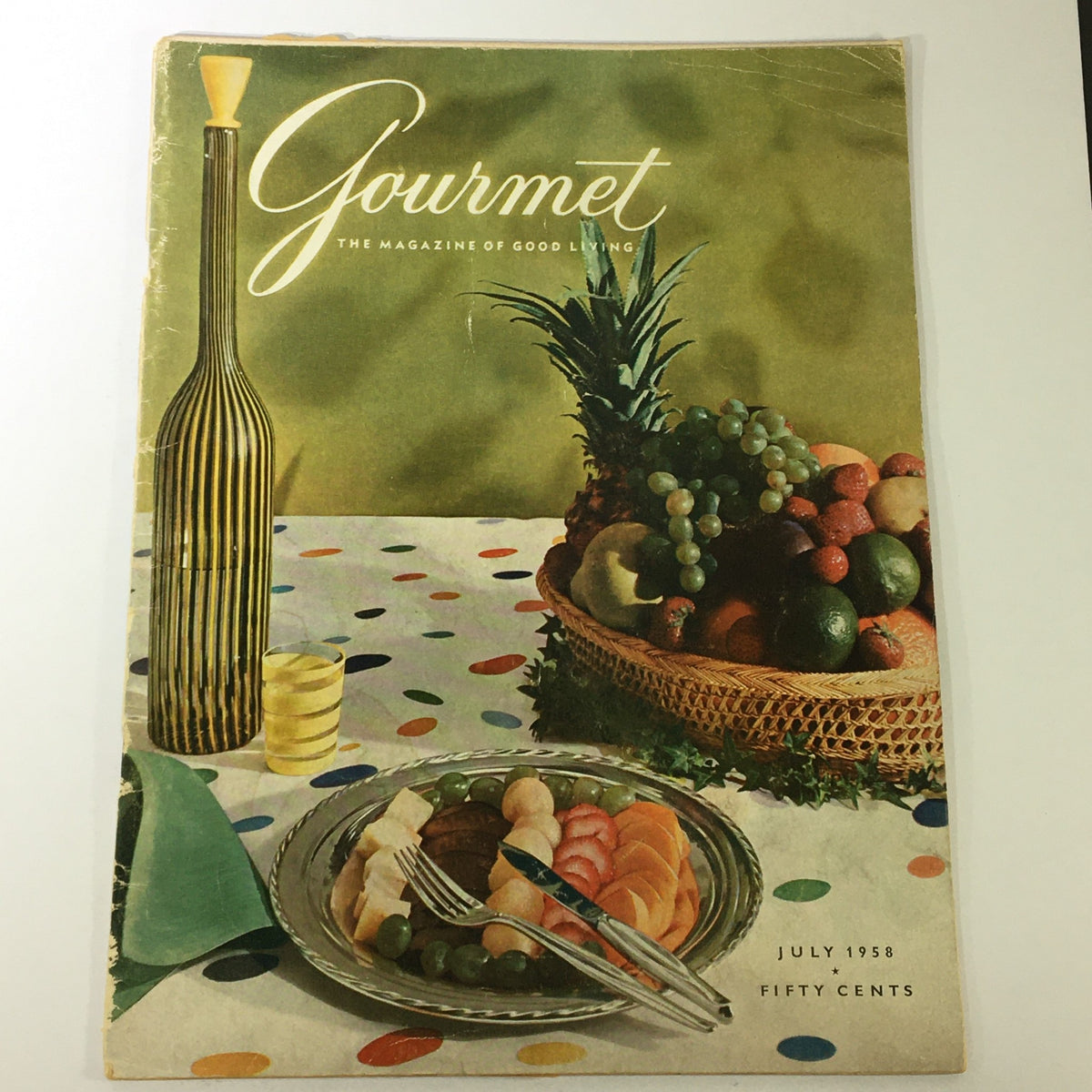 VTG Gourmet: The Magazine of Good Living July 1958 cover featuring a vibrant dining setting with a plate of assorted fruits and a decorative bottle, showcasing mid-century design aesthetics.