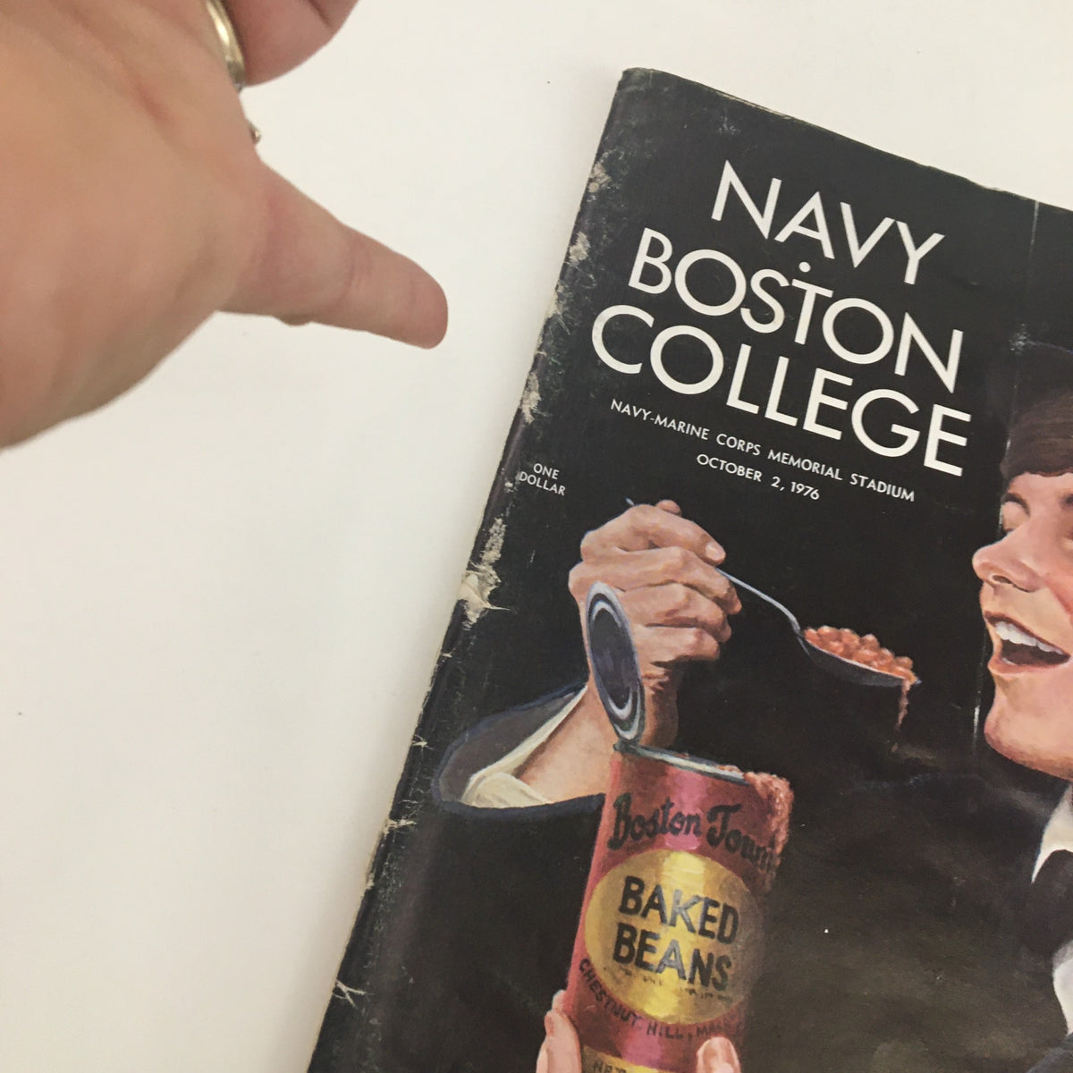 Close-up view of the 1976 Navy Boston College program cover with a sailor holding a Boston Baked Beans can, highlighting minor wear on the program.