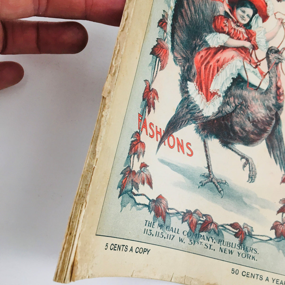 Side view of the cover of VTG McCall's Magazine November 1904, displaying the intricate illustration of a girl on a turkey with the word 'Fashions' highlighted.