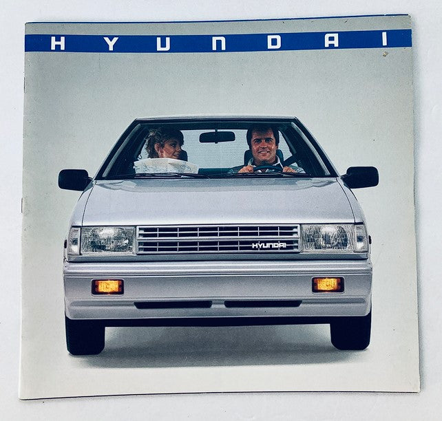 1980s Hyundai Dealer Showroom Sales Brochure featuring a silver Hyundai car with a couple inside, showcasing the vehicle's design and dealership promotional material.