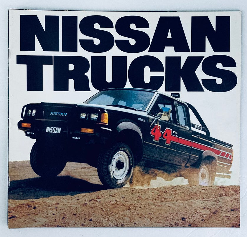 1984 Nissan Trucks Dealer Showroom Sales Brochure Guide Catalog featuring a black Nissan truck in action on rugged terrain, showcasing the brand's iconic styling and durability.