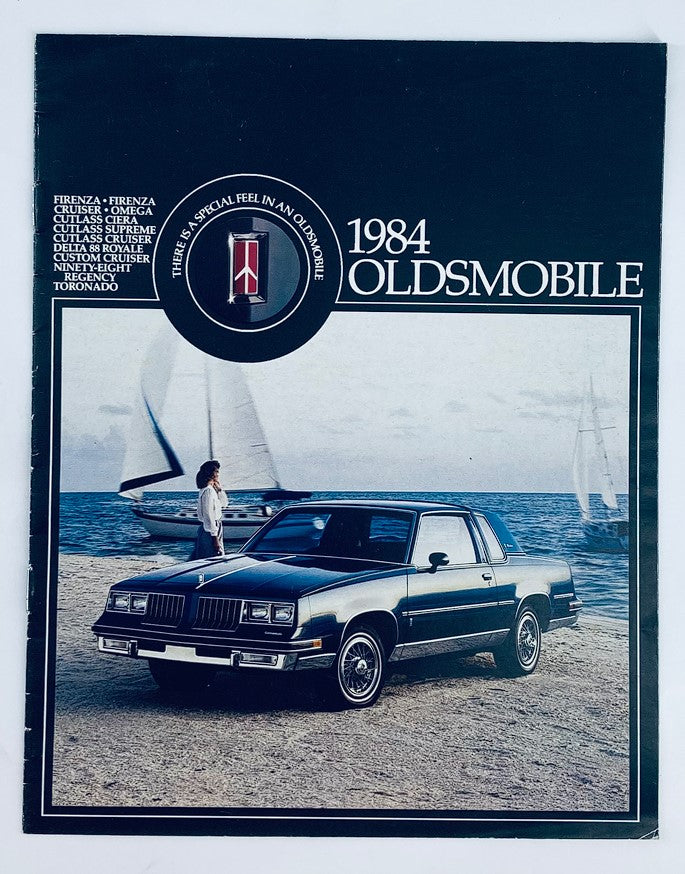 1984 Oldsmobile Firenza Cruiser Dealer Showroom Sales Brochure Guide Catalog showcasing the car parked near the beach with a person beside it, featuring the iconic Oldsmobile logo.
