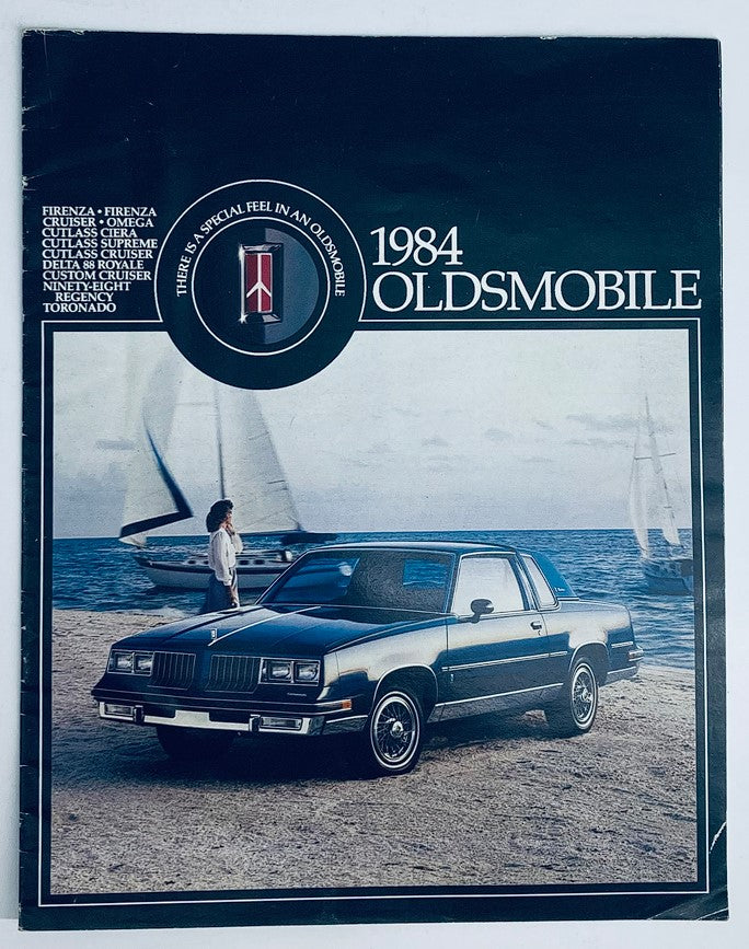 1984 Oldsmobile Firenza Dealer Showroom Sales Brochure featuring a stylish black Oldsmobile parked near the beach with sailboats in the background.