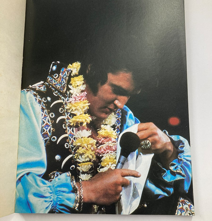 VTG 1979 Elvis in Concert by John Reggero capturing Elvis Presley wearing a floral lei and examining a microphone during a performance, highlighting his iconic presence.