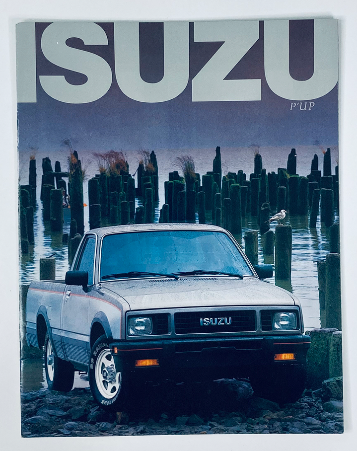 1987 Isuzu Pickup Dealer Showroom Sales Brochure Guide Catalog featuring a classic Isuzu Pup truck positioned near a serene water body, showcasing the vehicle's design against a backdrop of wooden posts and a calm atmosphere.