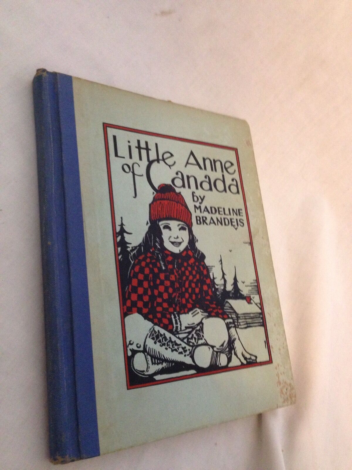 Front cover of the vintage book 'Little Anne of Canada' by Madeline Brandeis, published in 1931, featuring a young girl in a red checkered shirt and a winter hat, set against a backdrop of trees and a cozy cabin.