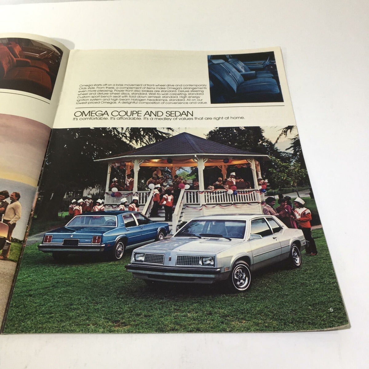 Page from the 1983 Oldsmobile Firenza 'S' Coupe and 4-Door Sedan brochure displaying two cars, a blue and a silver model, parked near an outdoor gazebo with a crowd.