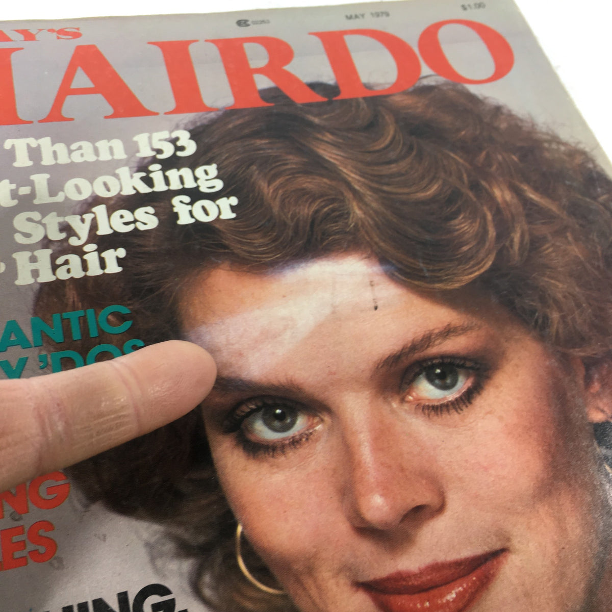 Close-up of the cover of VTG Today's Hairdo Magazine: May 1979 - Pretty Spring Styles with a finger pointing at the featured hairstyle, highlighting the magazine's emphasis on spring hair trends.