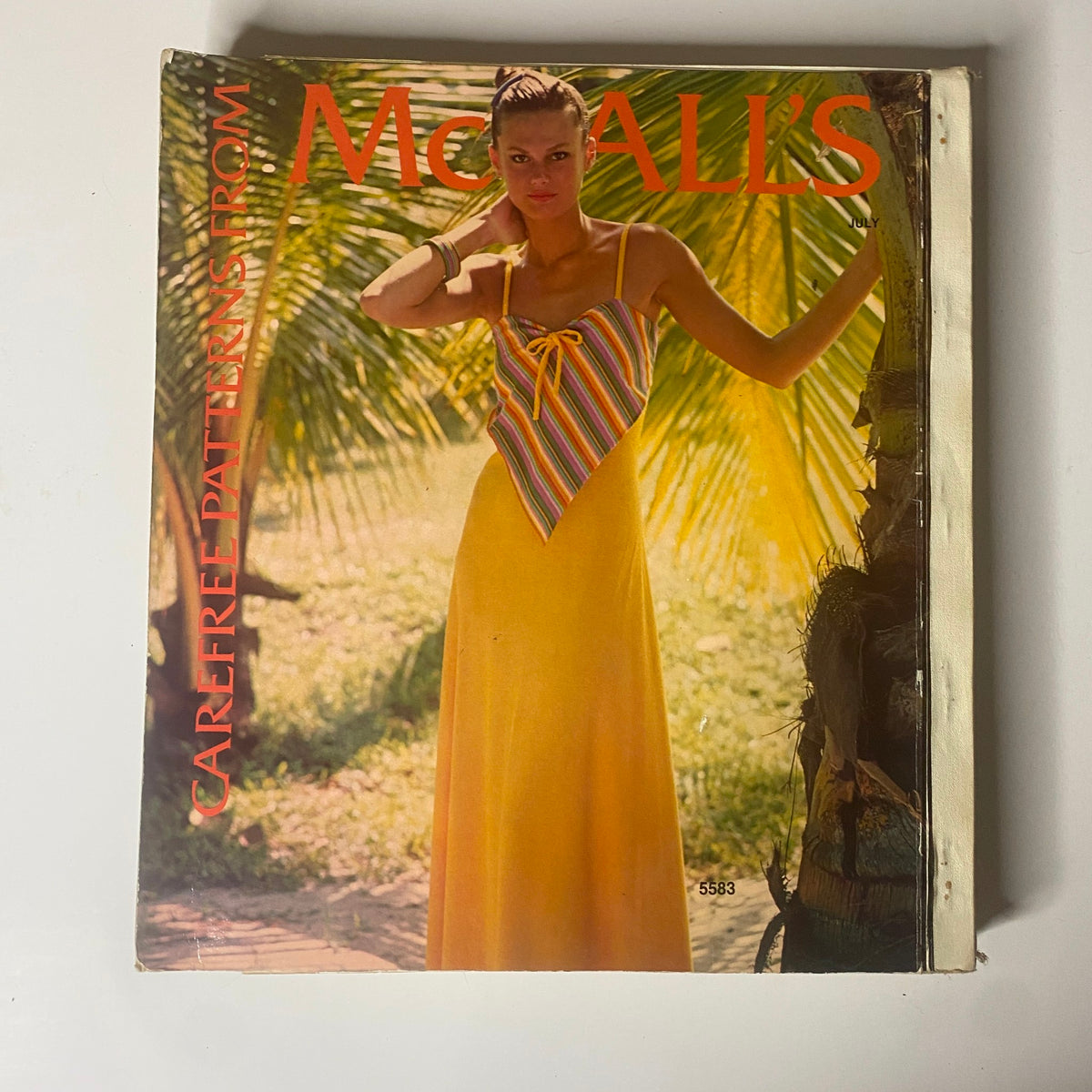 1977 July Carefree Patterns McCall's Summer Catalog featuring a model in a vibrant yellow summer dress with a striped top. The backdrop showcases lush greenery, embodying a relaxed summer vibe.