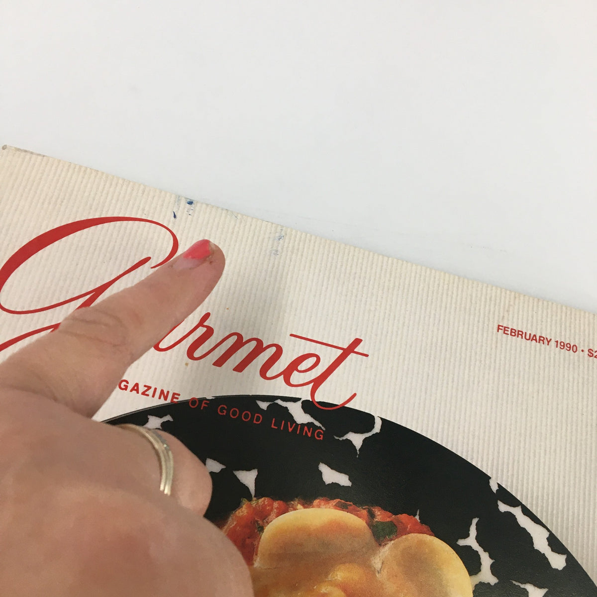Close-up of Gourmet Magazine February 1990 showing a finger pointing to minor wear on the magazine cover, highlighting the condition of the publication.