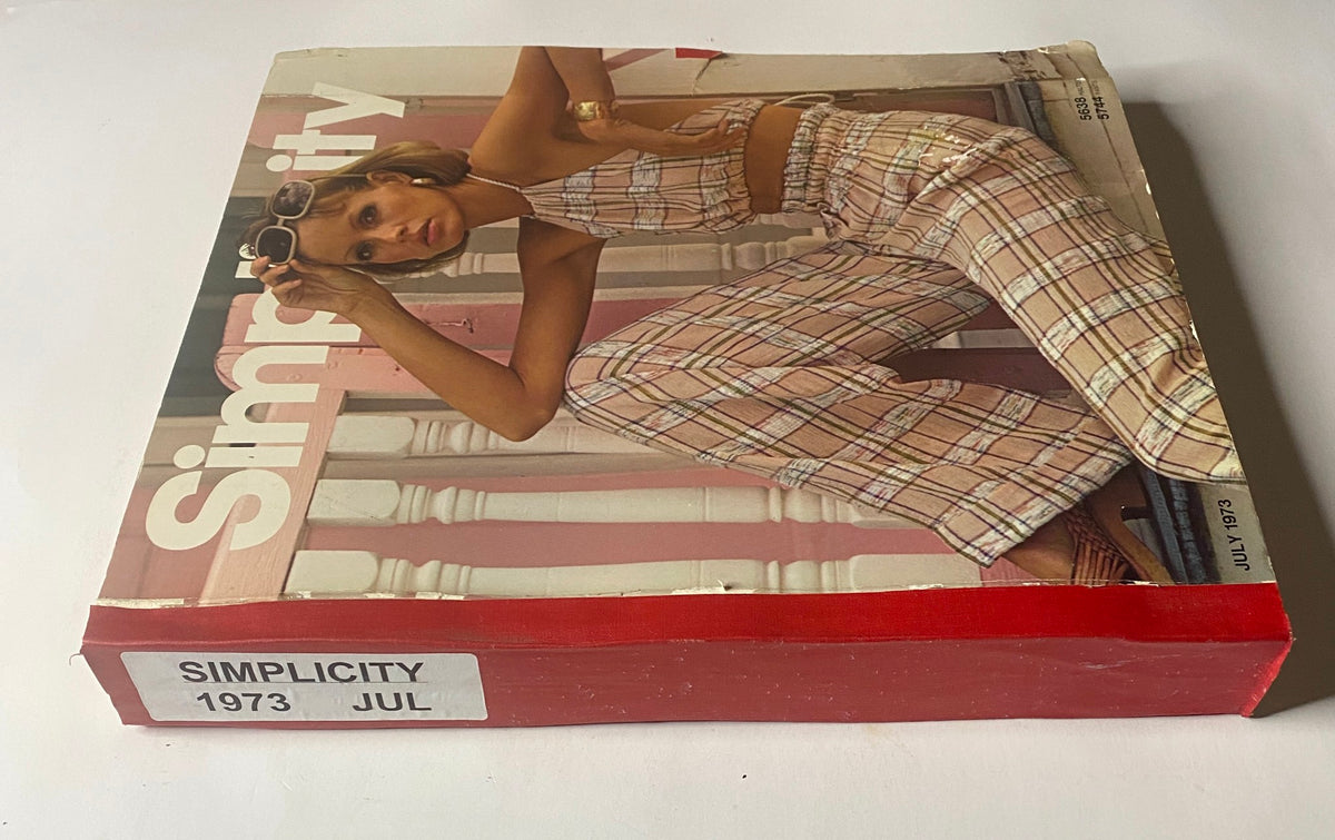 1973 July Simplicity Catalog displayed with a young woman in a plaid outfit, showcasing summer fashion trends and sewing patterns from the mid-1970s.