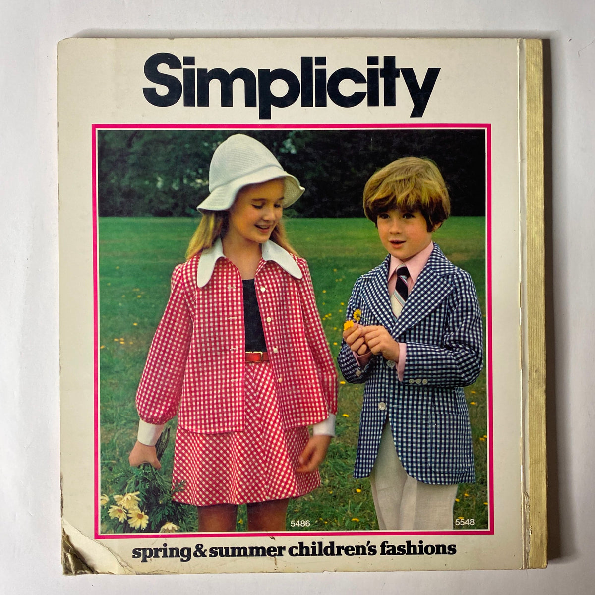 1973 Simplicity Spring and Summer Catalog showcasing children's fashions. The cover features a girl in a pink checkered dress with a matching coat and a boy in a blue checkered suit. This visually striking design captures the playful essence of 1970s fashion for kids.