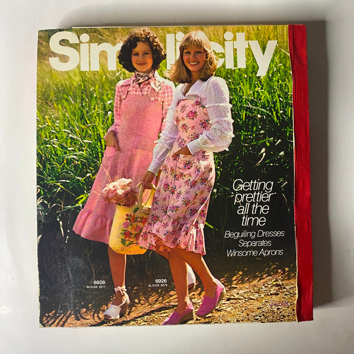 1970s women's fashion displayed in the 1975 June Simplicity Catalog. Two models wearing charming floral dresses and stylish outfits in a verdant outdoor setting, emphasizing vintage style, with the title '1975 June Simplicity Catalog Sewing Pattern Giant Store Counter Book Fashion'.