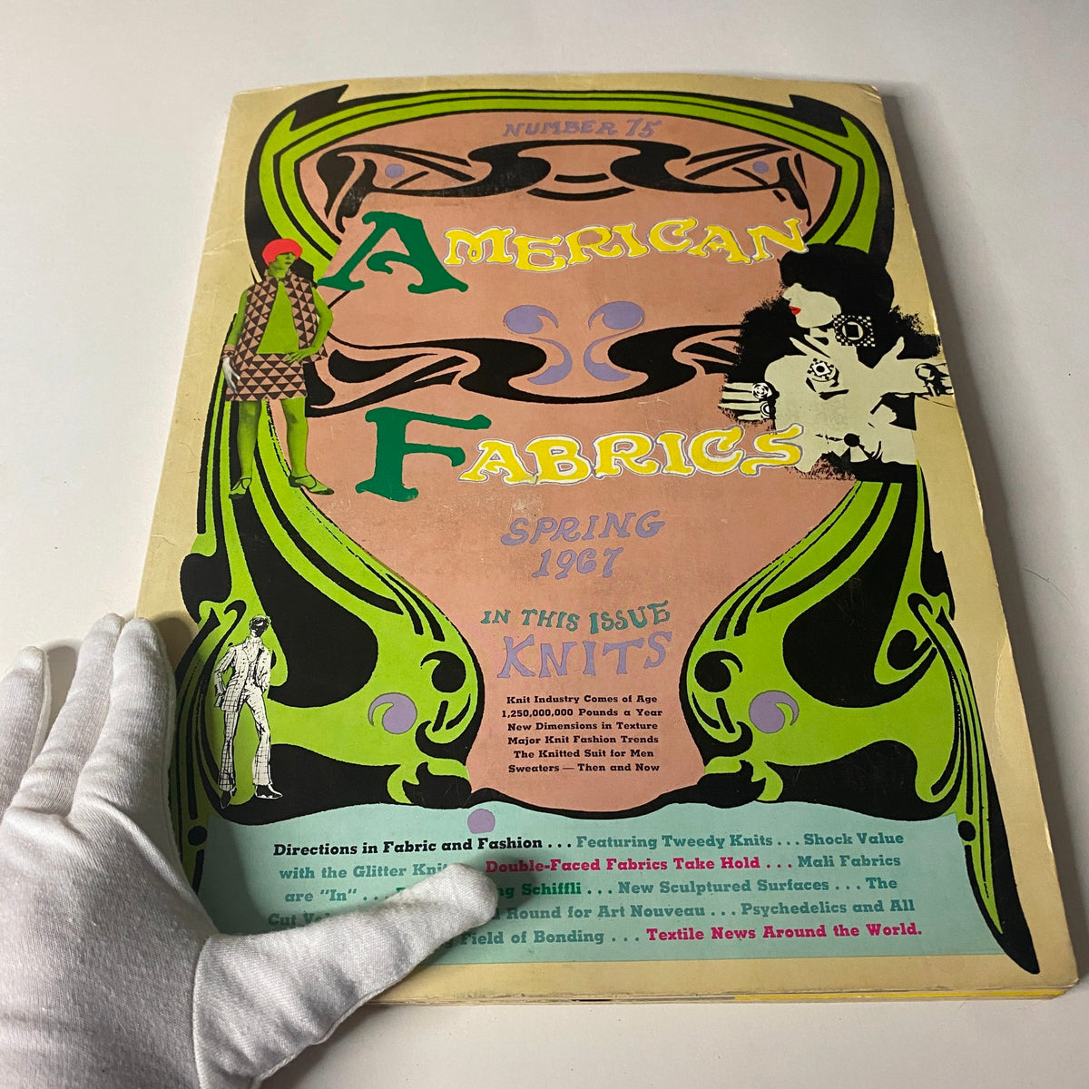 Image of the 1967 American Fabric No. 75 Spring Catalog being held, displaying its eye-catching cover with fashion figures and intricate graphics related to textile design.