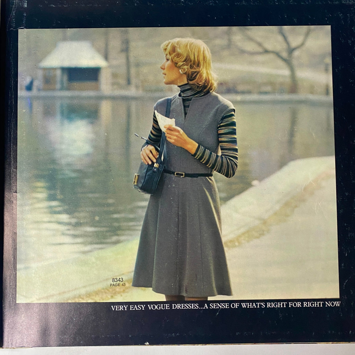 1972 Vogue Patterns September Catalog cover featuring a stylish woman with a chic haircut, wearing a grey sleeveless dress and striped long-sleeve shirt, standing by a serene water body.
