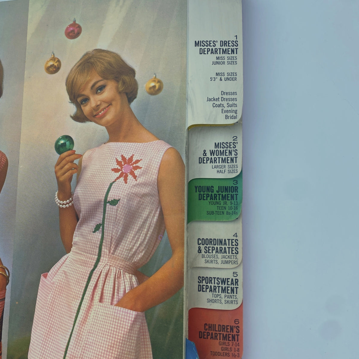 Close-up from the 1961 December Butterick Holiday Catalog highlighting a model in a pink dress adorned with floral embroidery, surrounded by holiday ornaments.