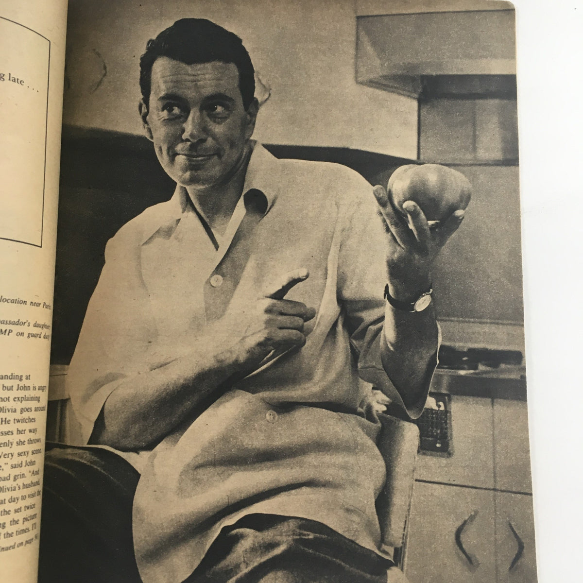 Black and white image featuring a male celebrity holding an apple in a kitchen setting, from Hollywood Stars Magazine October 1956, illustrating casual celebrity moments from the era.