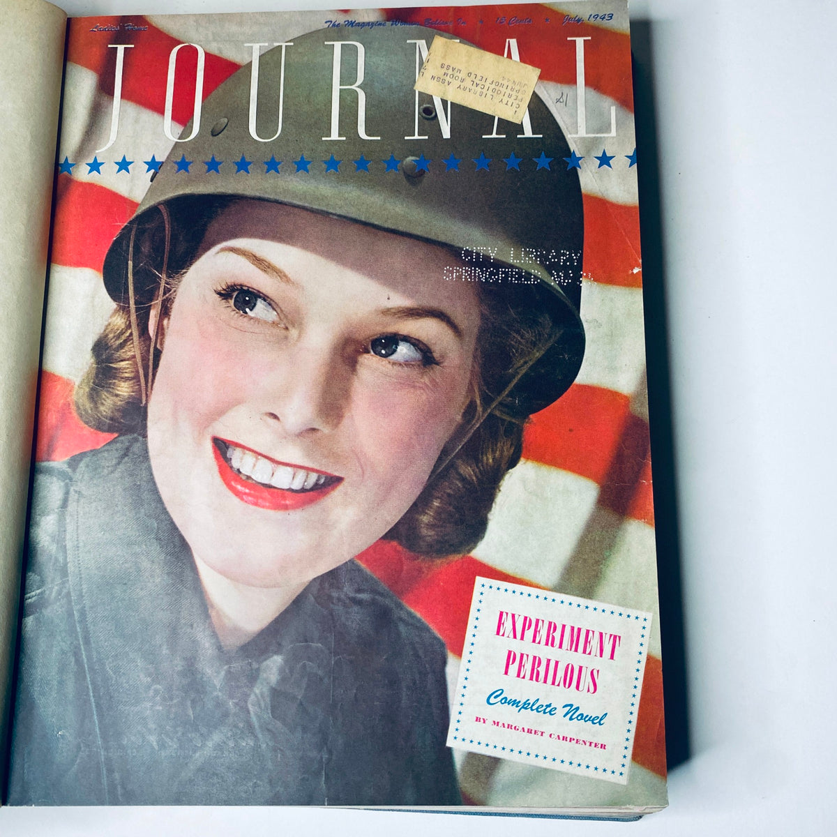 Cover of Ladies' Home Journal, July 1943 edition, featuring a smiling woman in a military helmet with an American flag backdrop. Magazine highlights include 'Experiment Perilous' complete novel.