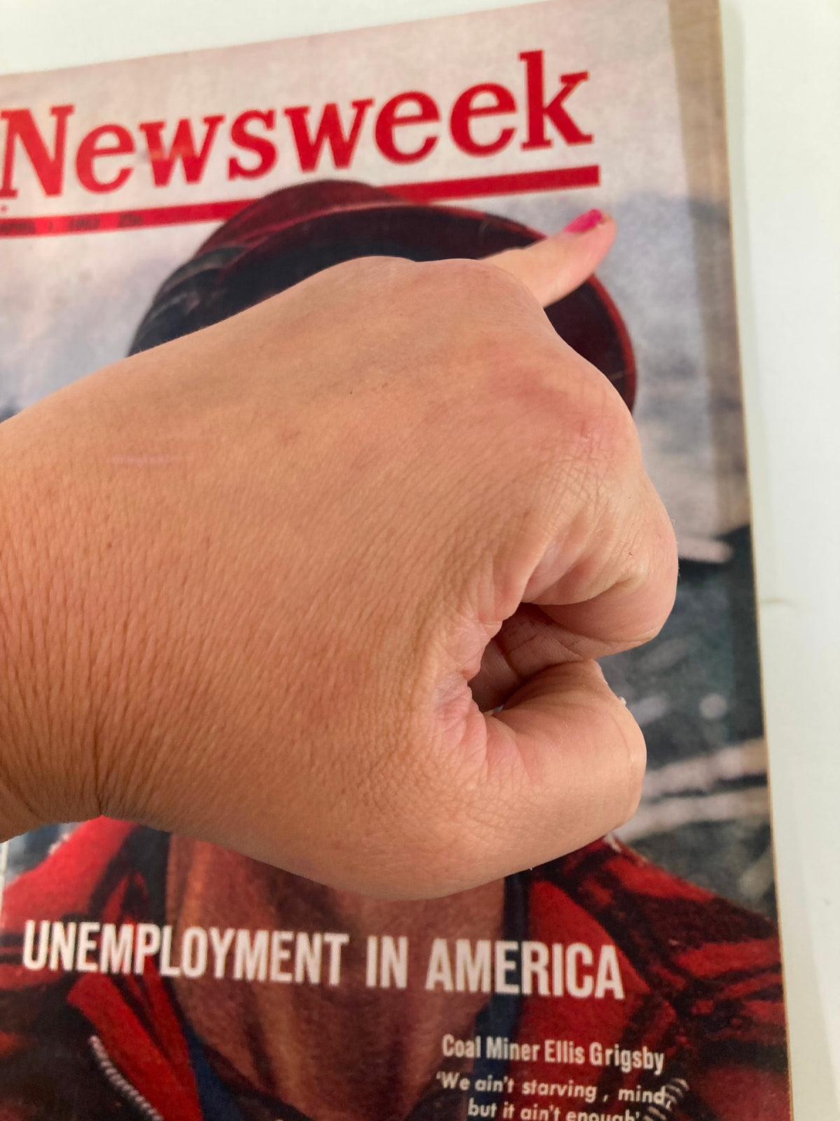 Close-up of a hand pointing to the title 'Newsweek' on the vintage magazine cover from April 1, 1963, featuring a story about 'Unemployment in America' by Miner Ellis Grigsby. The hand is indicating the magazine's prominent header, highlighting its historical significance.