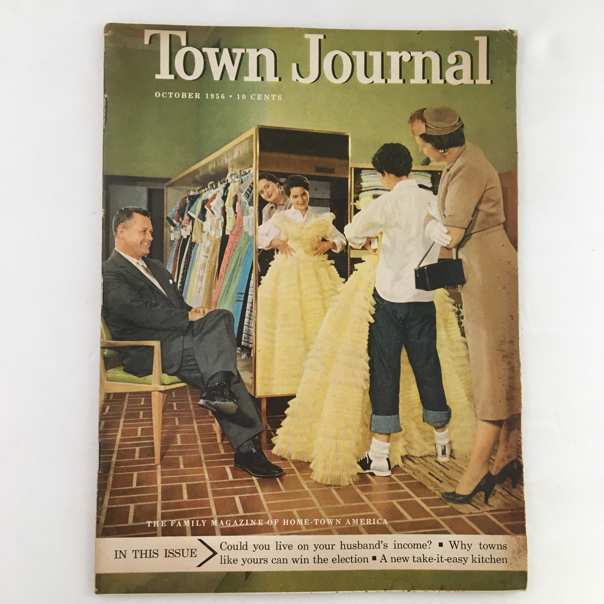 Cover of Town Journal Magazine October 1956, featuring the cover story 'Could You Live On Your Husband's Income'. The image showcases a vintage scene with a man seated, observing women in elegant dresses at a clothing store.