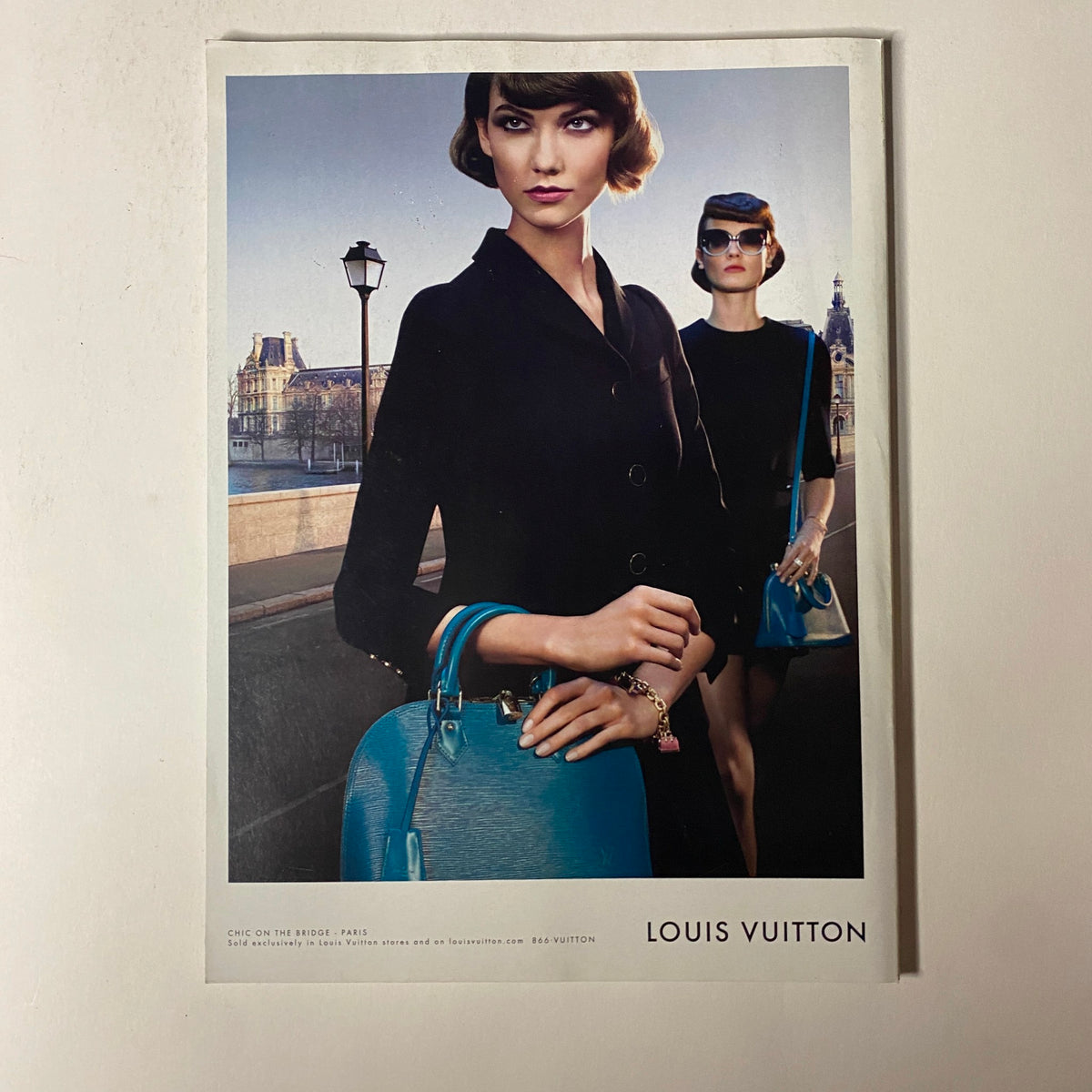Fashion advertisement from Vogue Magazine July 2013 promoting Louis Vuitton, featuring two models dressed in elegant black attire with striking blue accessories against a scenic background.