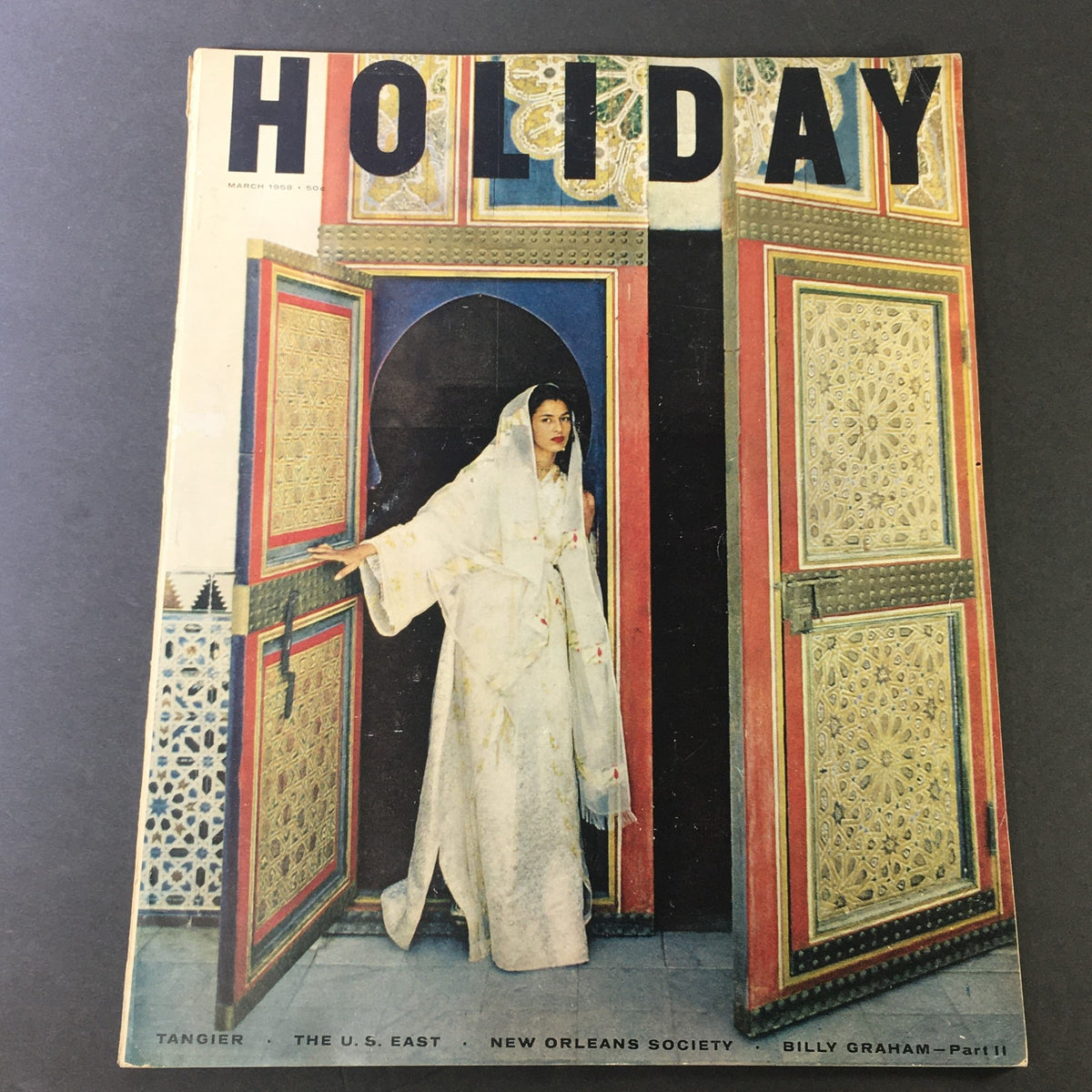 Cover of VTG Holiday Magazine March 1958 featuring a woman adorned in traditional attire standing at an ornate door, highlighting Tangier, the U.S. East, and Billy Graham Part II.