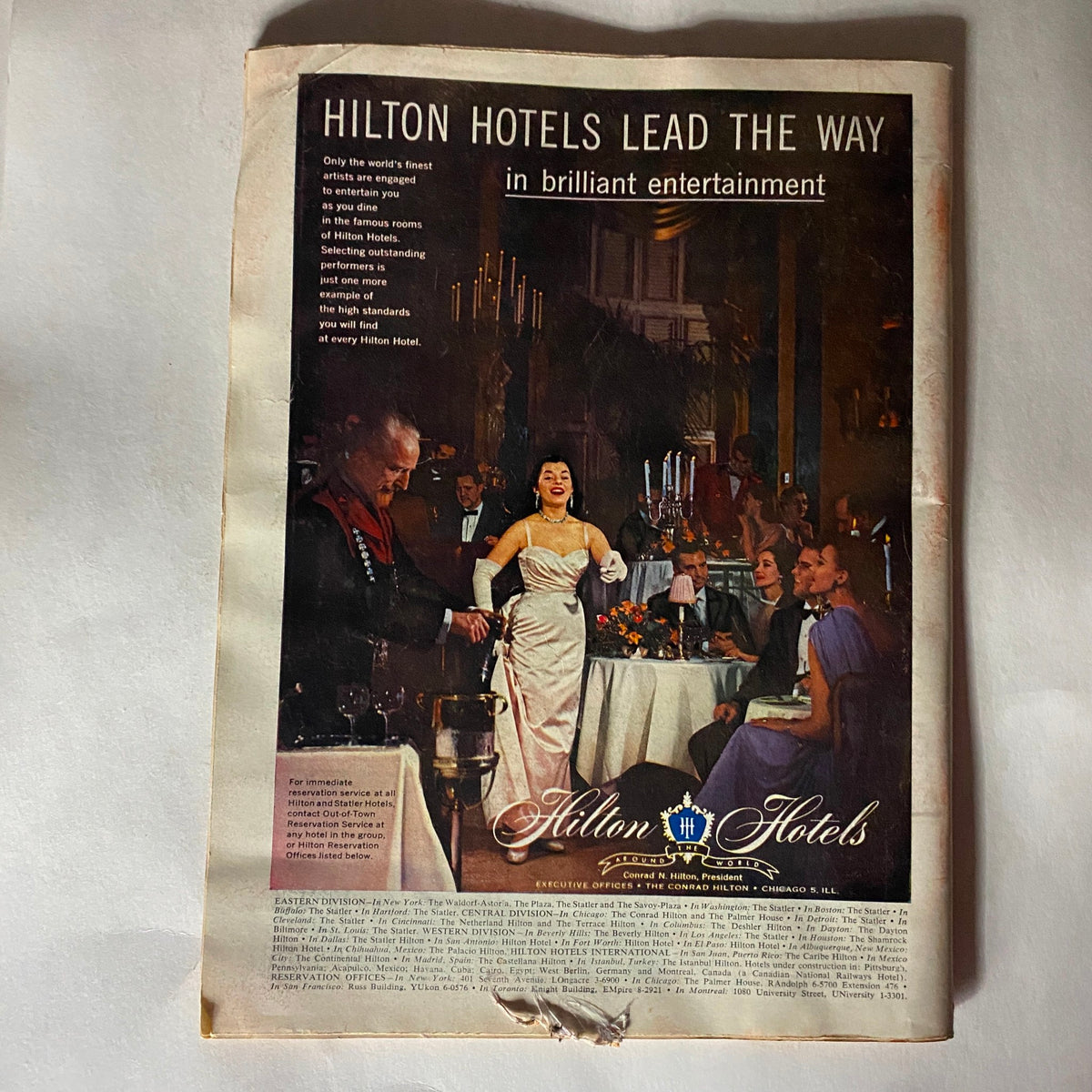 Advertisement from VTG Time Magazine March 25, 1957 Vol 69 No. 12 for Hilton Hotels, showcasing a glamorous dining scene with elegantly dressed guests and a performer. The ad highlights Hilton's commitment to fine entertainment.