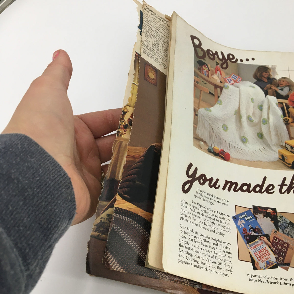 Image of a finger holding a page from McCall's Needlework & Crafts Magazine February 1983, depicting a cozy home scene and promoting handcrafted items from Boye.