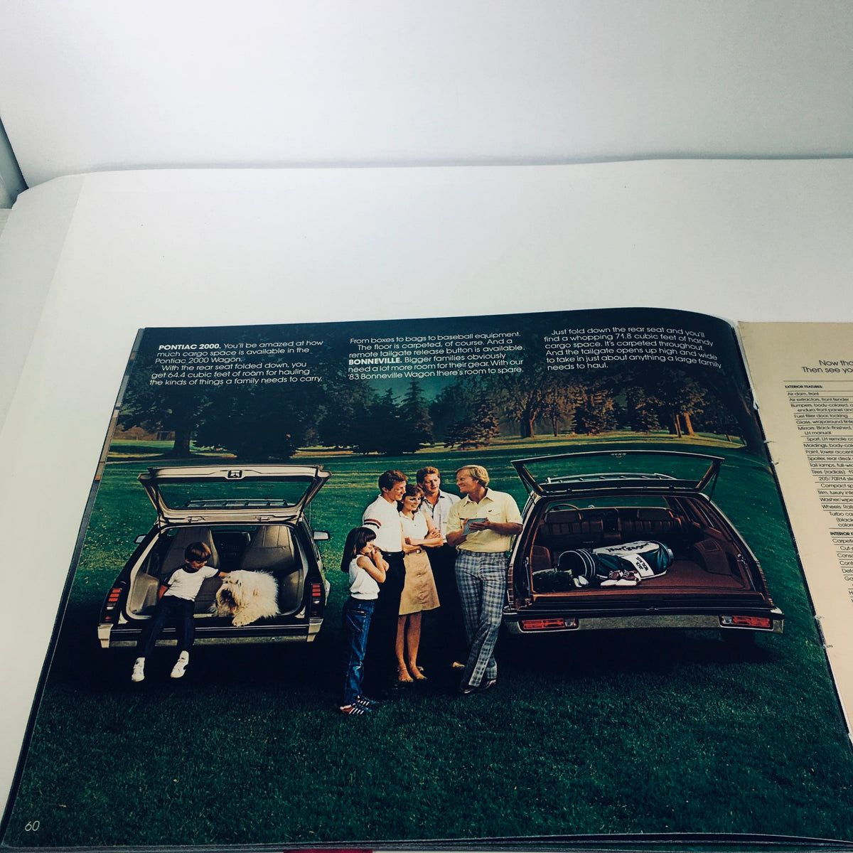 Promotional image from the 1983 Pontiac 2000 5.8 Liter V-8 Engine Grand Prix Car Sale Brochure featuring a family interacting around two cars in a park setting.